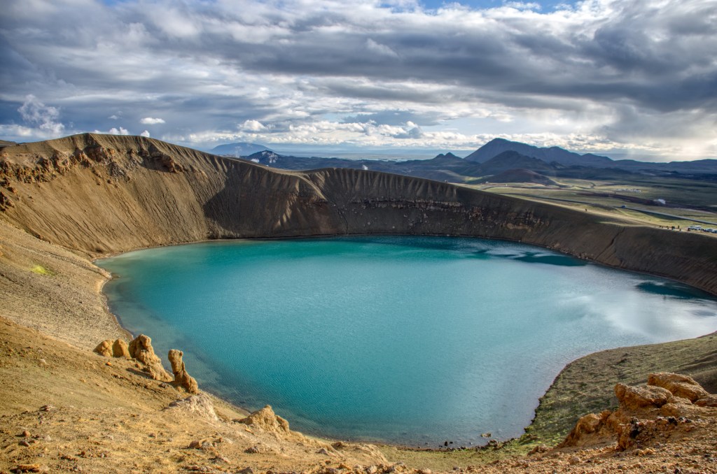Large, turquoise blue lake in a rocky caldera with mountains in the distance under cloudy skies