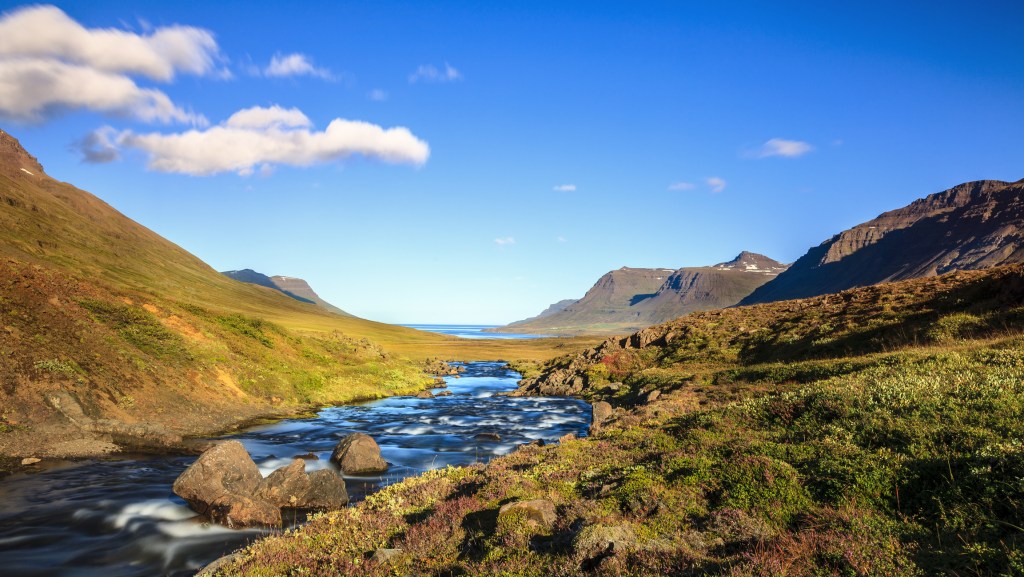 A stream running through a lush valley under blue sky.