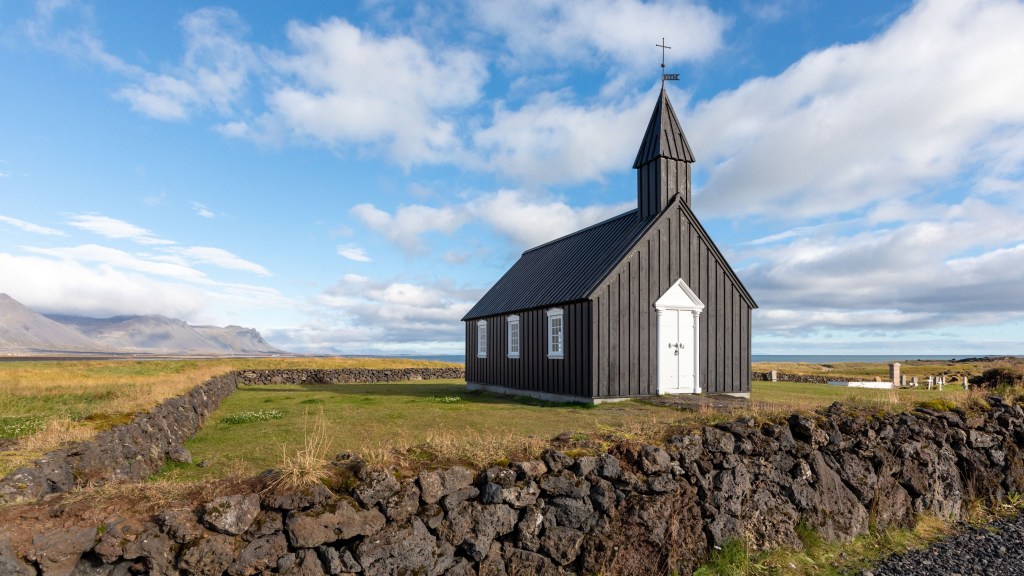 A small black wooden church surrounded by a low stone wall under a blue cloudy sky