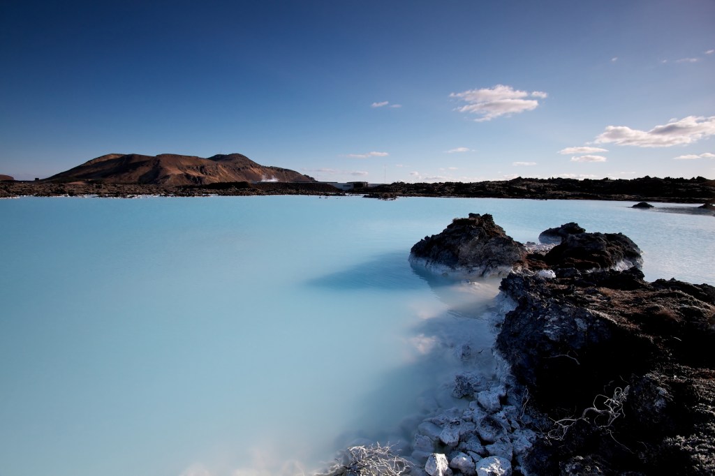 Calm, milky blue waters and dark rocks stretching out toward low hills in the distance