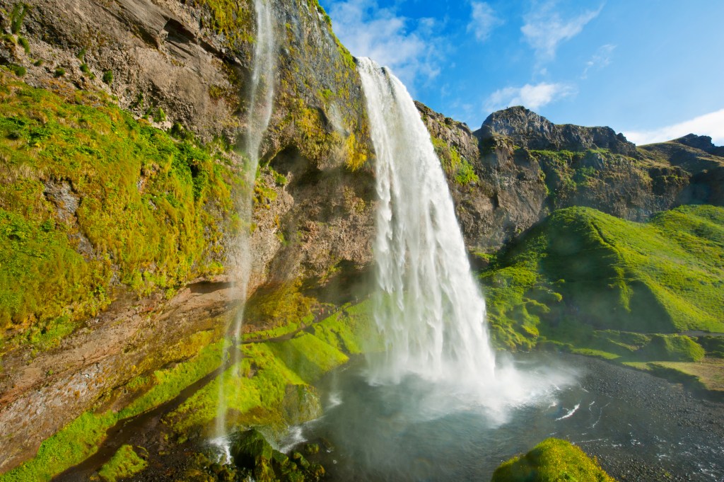 Waterfall rushing over the side of stunning mossy green cliff under bright blue sky.