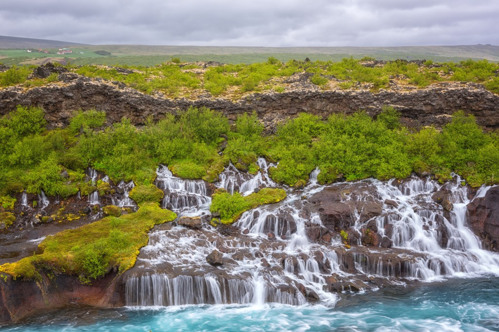A swath of delicate cascading waterfalls pouring down over rocks on lush, bright green hillside.